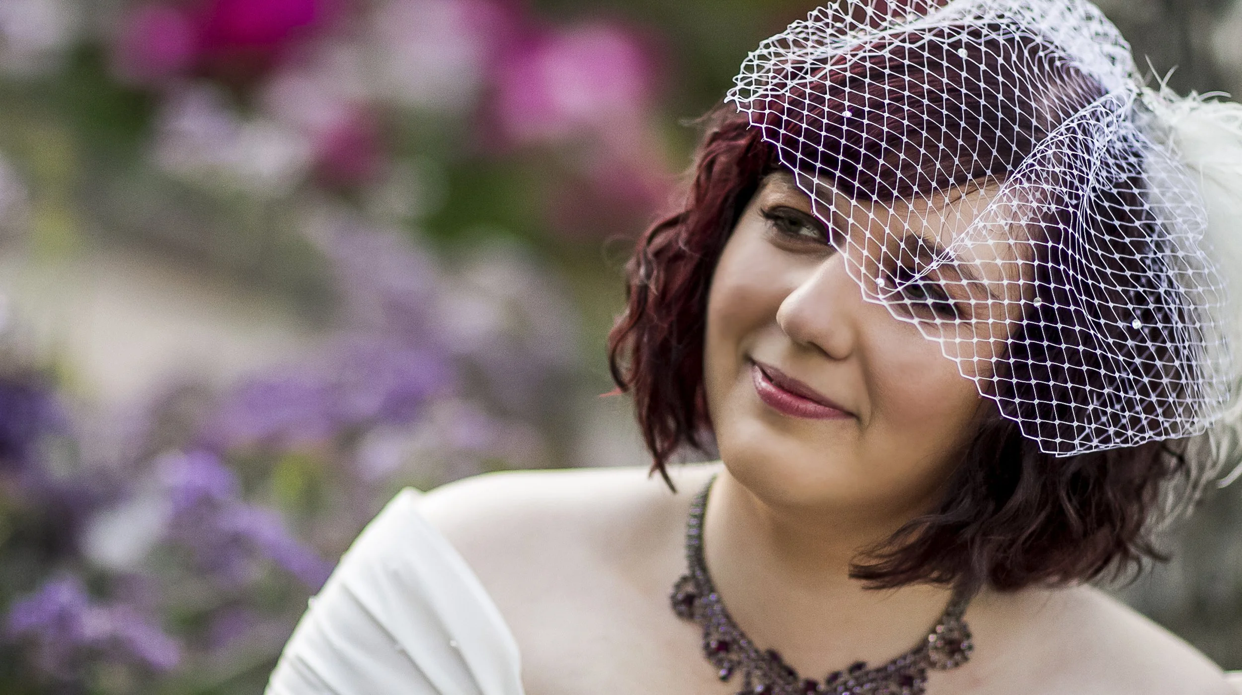 A woman with shoulder-length curly hair, wearing a purple fascinator with netting, a white dress, and a beaded necklace, smiling outdoors with blurred purple flowers in the background.