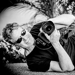 Man lying on grass, holding a camera, smiling, wearing sunglasses, outdoors with palm trees.