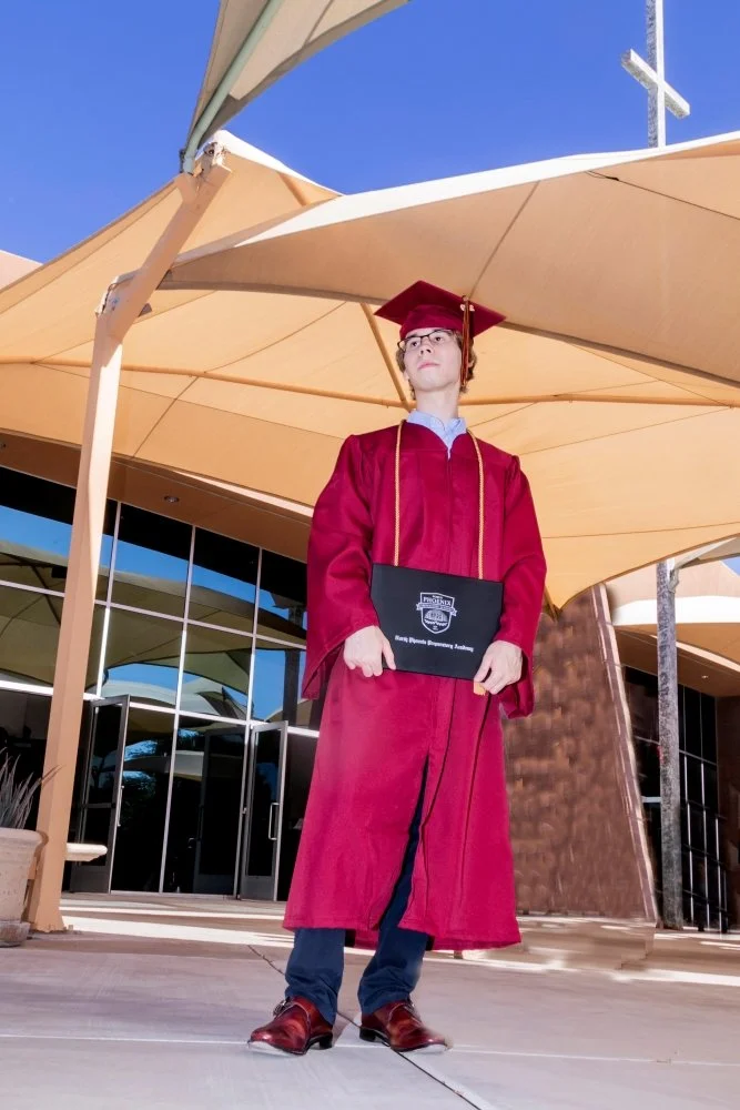 A young man in a red graduation gown and cap holding a diploma stands outside under a large beige umbrella, in front of a building with glass doors.