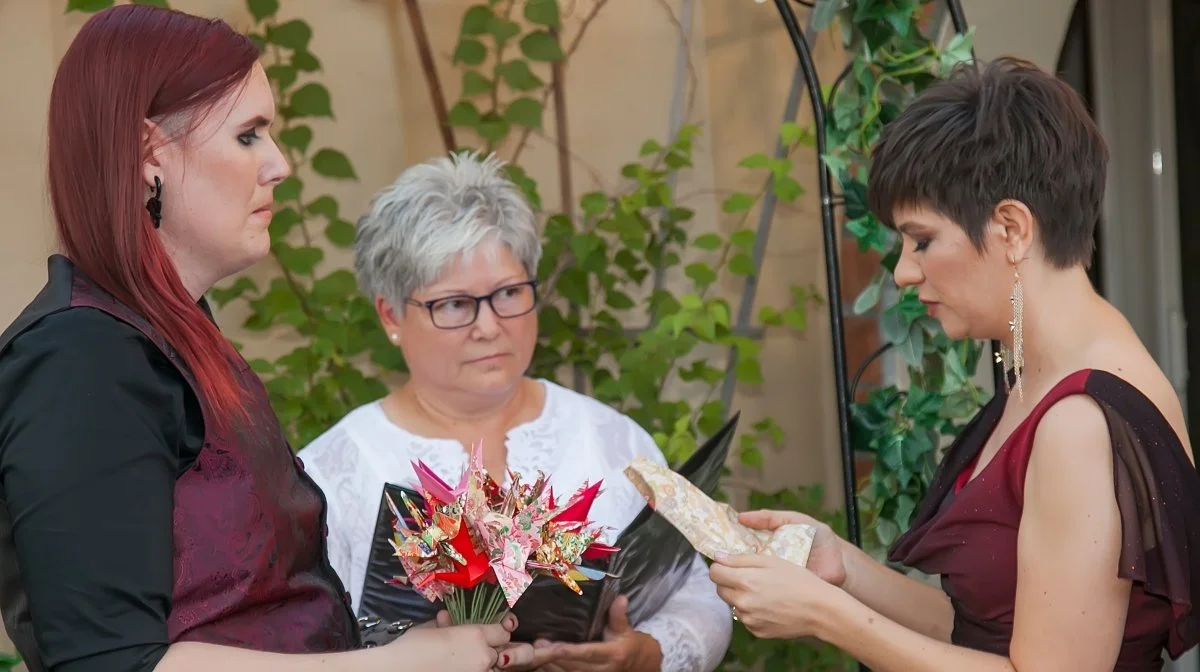 Three women at a celebration, one holding a bouquet of origami flowers, another reading a paper, with greenery in the background.