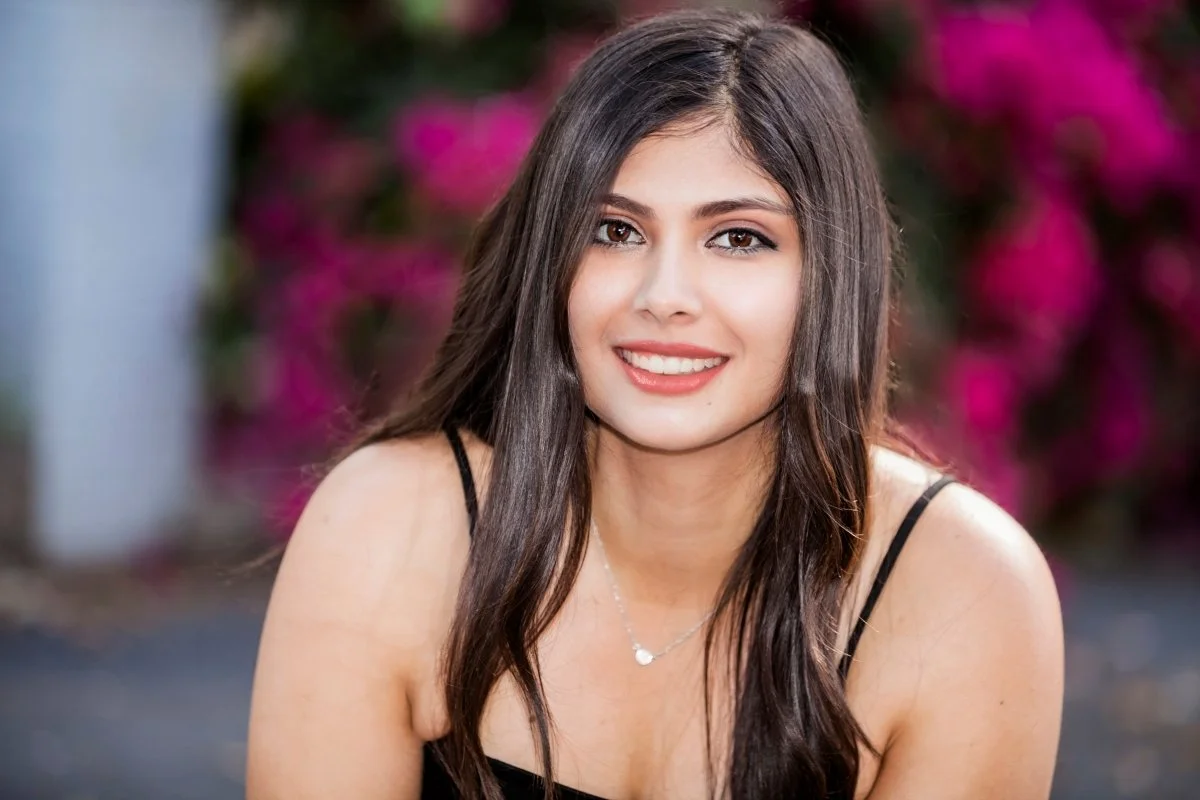 Young woman with long dark hair wearing a black spaghetti strap top and a small necklace, smiling outdoors with pink flowers in the background.
