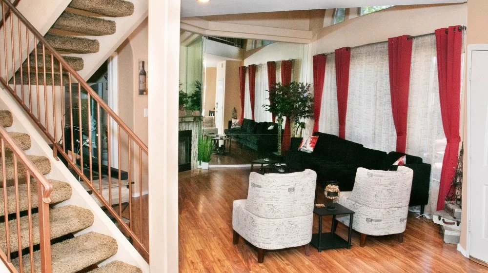 Living room with wooden flooring, black sofa, white armchairs, red curtains, and large windows.