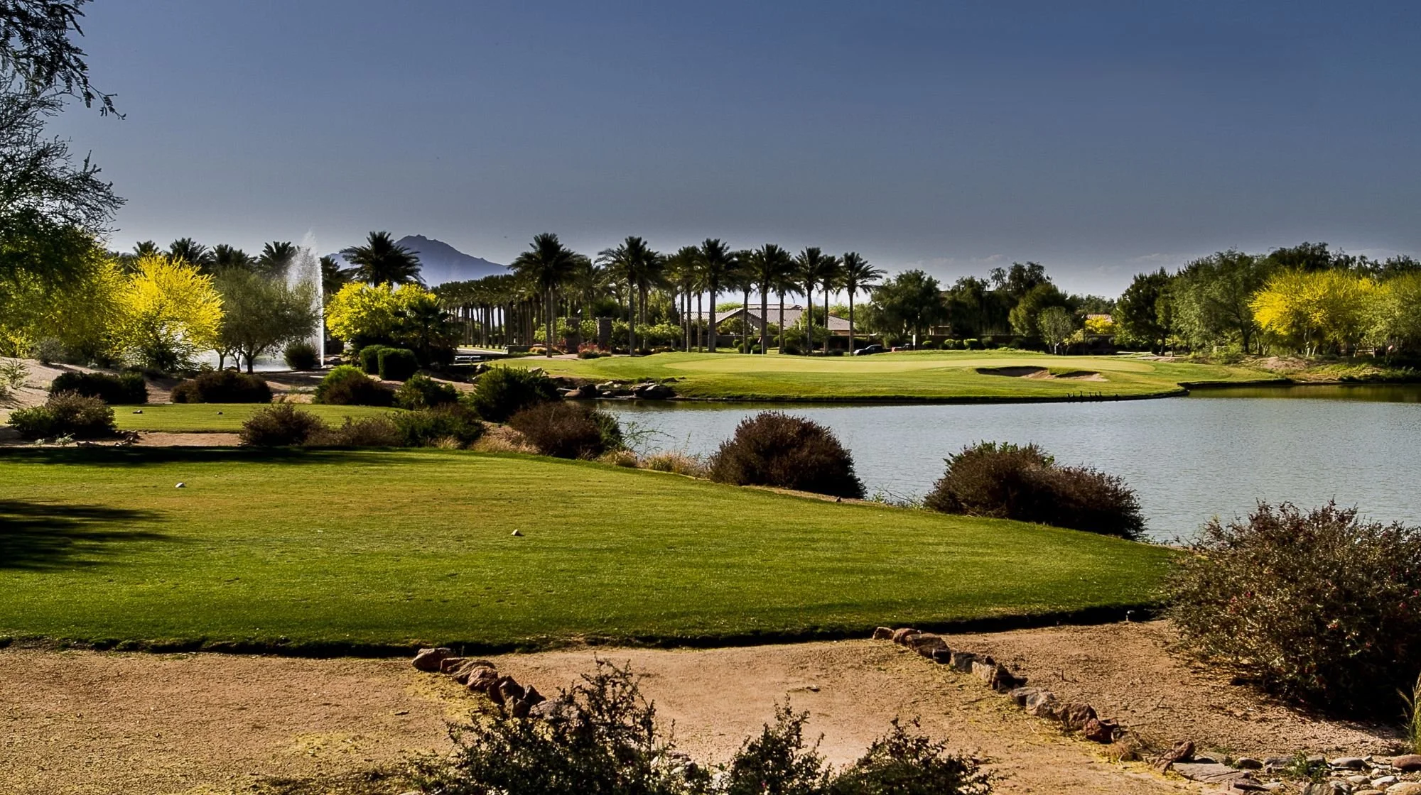 A golf course with a water hazard, lush green grass, trees, and palm trees, mountains in the background, and a fountain in the water, under a clear blue sky.