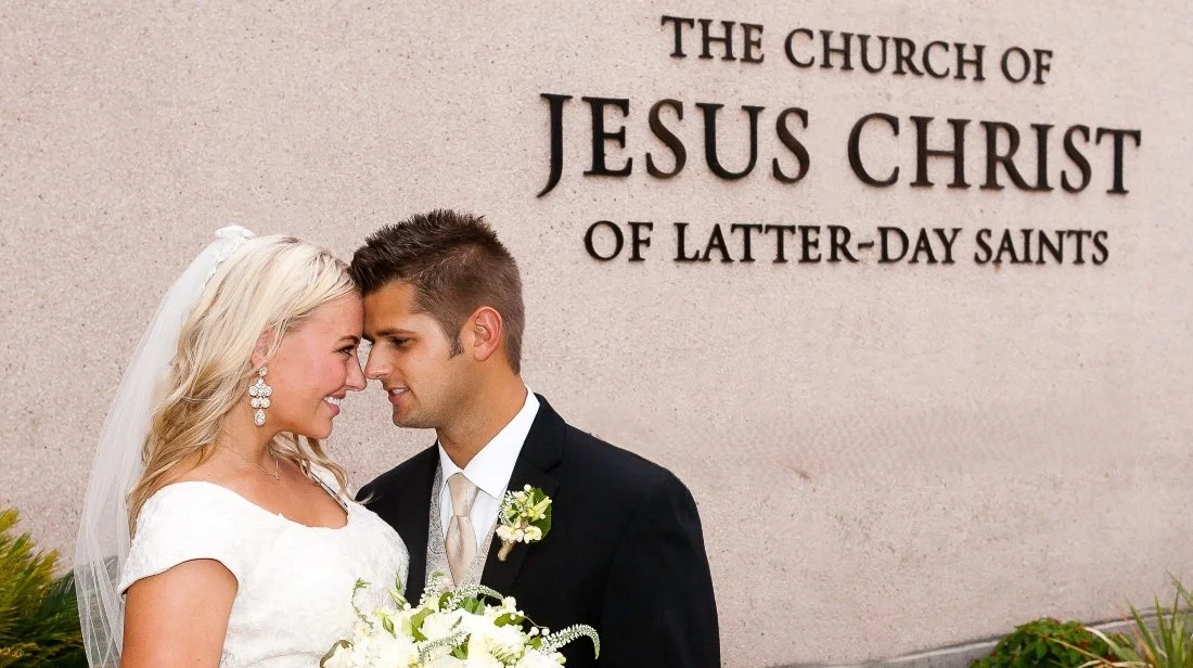 A bride and groom at their wedding standing close together with foreheads touching outdoors in front of a wall with text that reads 'The Church of Jesus Christ of Latter-day Saints'.