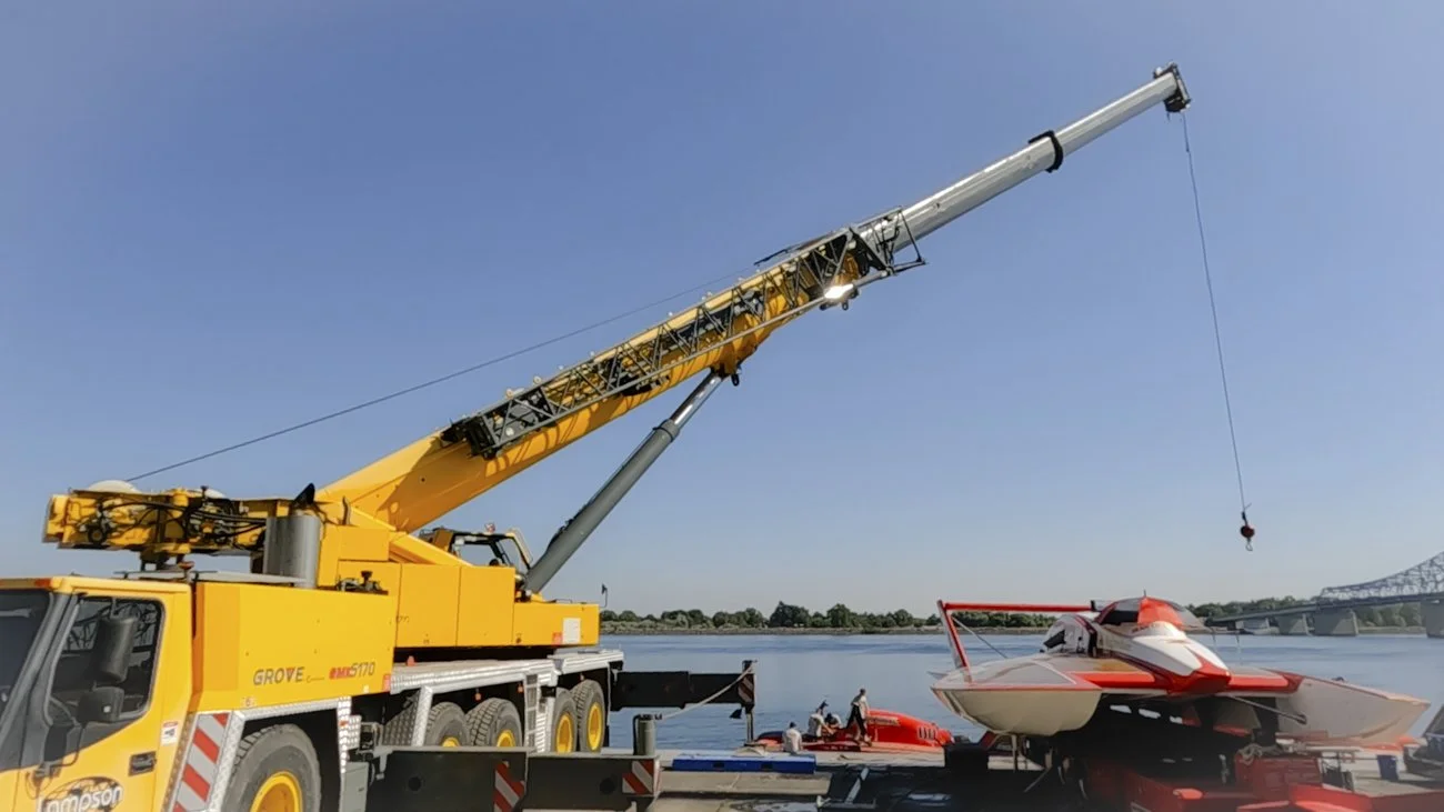 A large yellow crane lifting a red and white hydroplane boat near a body of water with people on small boats in the background.