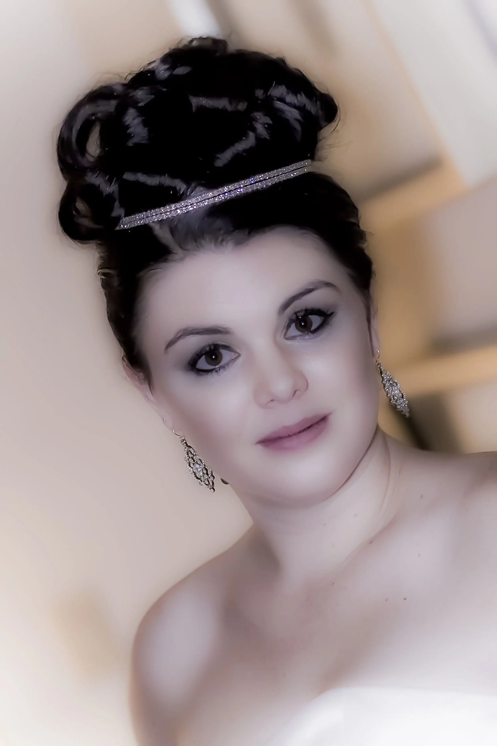 A woman with styled dark hair in an elegant updo, wearing sparkly earrings, with makeup, and wearing a strapless dress, standing indoors against a light-colored background.