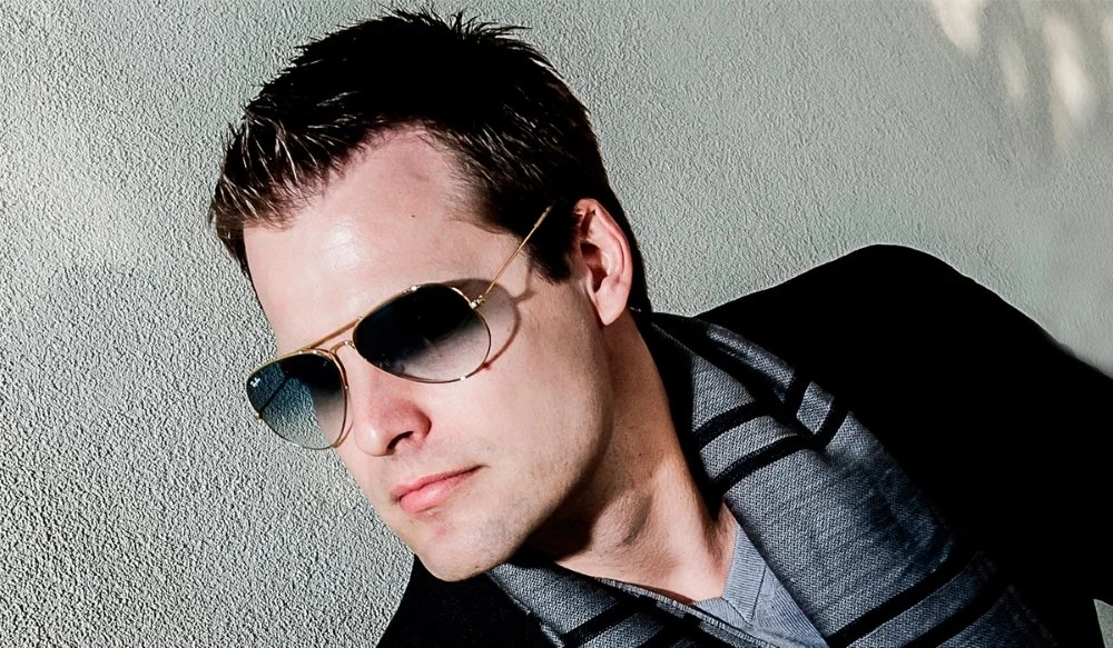 A man wearing sunglasses and a striped shirt with a dark jacket, looking down and to the side against a textured wall.