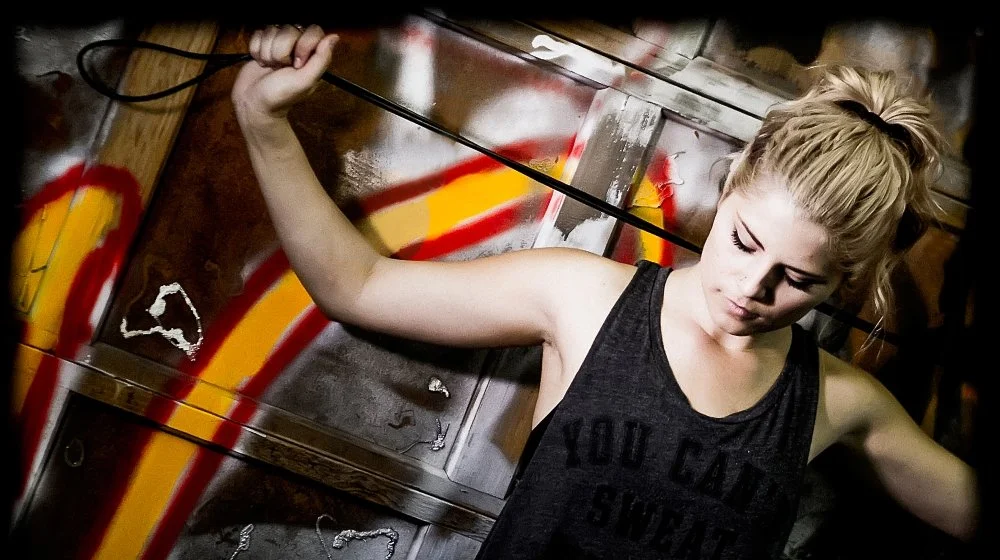 A young woman with blonde hair tied up in a messy ponytail, wearing a black sleeveless shirt, stands against a graffiti-covered metal surface. She holds a whip in her right hand, raised above her head, and appears to be in a determined or contemplati