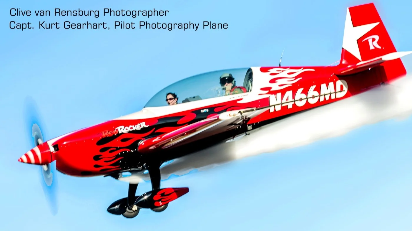 A red and black aerobatic airplane with flame decals flying in a clear blue sky. Two pilots are visible in the cockpit, wearing helmets and goggles. Text in the upper left corner credits Clive van Rensburg as the photographer and Captain Kurt Gearhar