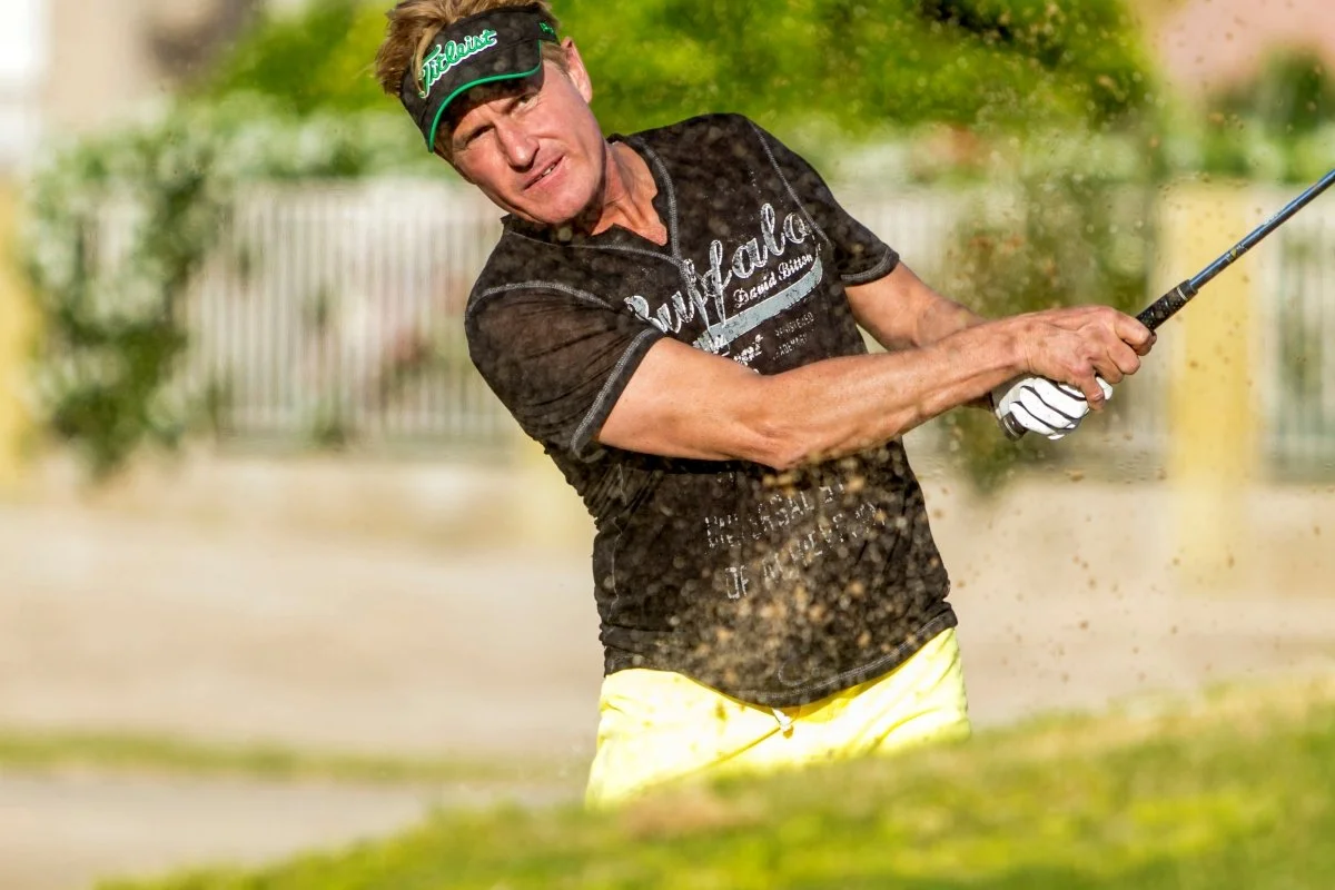 A man wearing a black t-shirt, yellow shorts, and a green visor is swinging a golf club, hitting a ball out of a sand trap on a golf course.