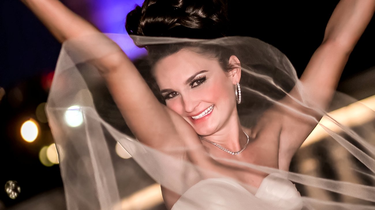 A woman smiling, dressed in wedding attire with a veil, with her arms raised, at night with blurred colorful lights in the background.