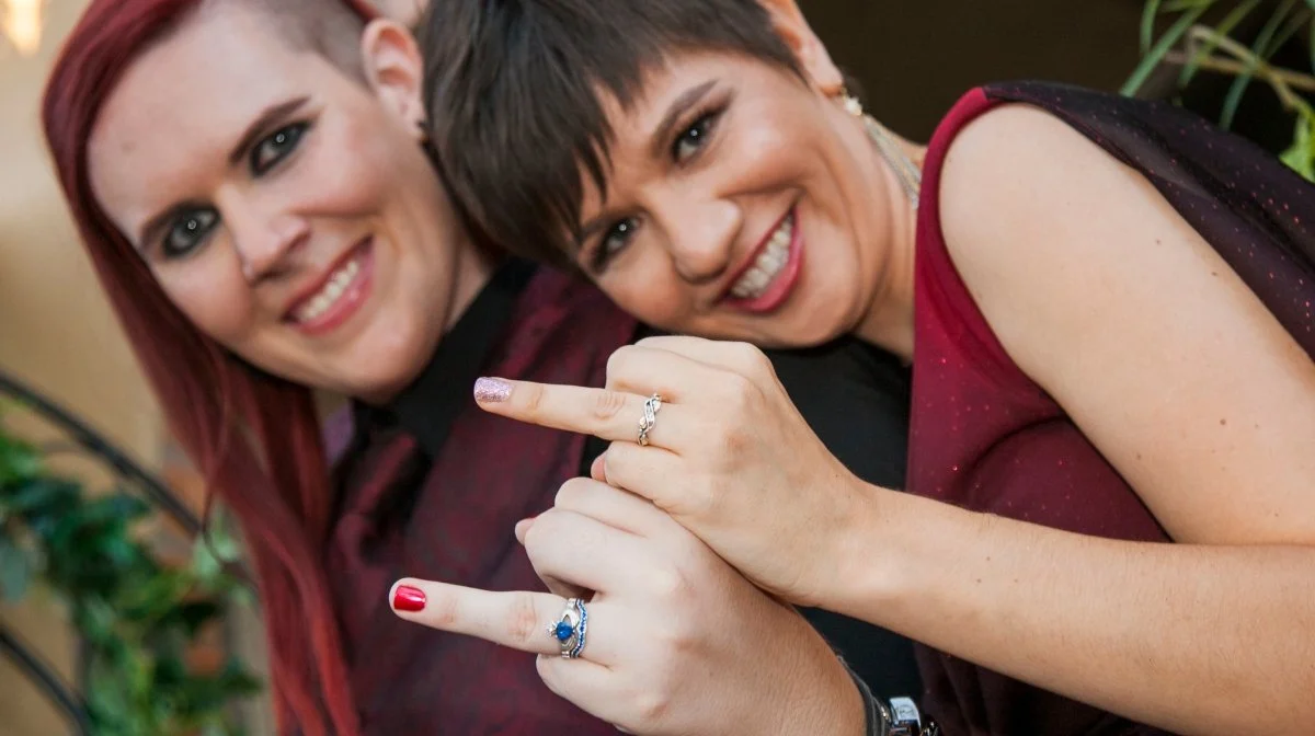 Two women smiling and showing off rings on their fingers, holding hands with interlocked fingers.