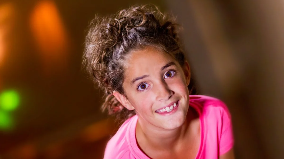A young girl with curly hair wearing a pink shirt, smiling and tilting her head at an angle, with a blurred background with warm lighting.