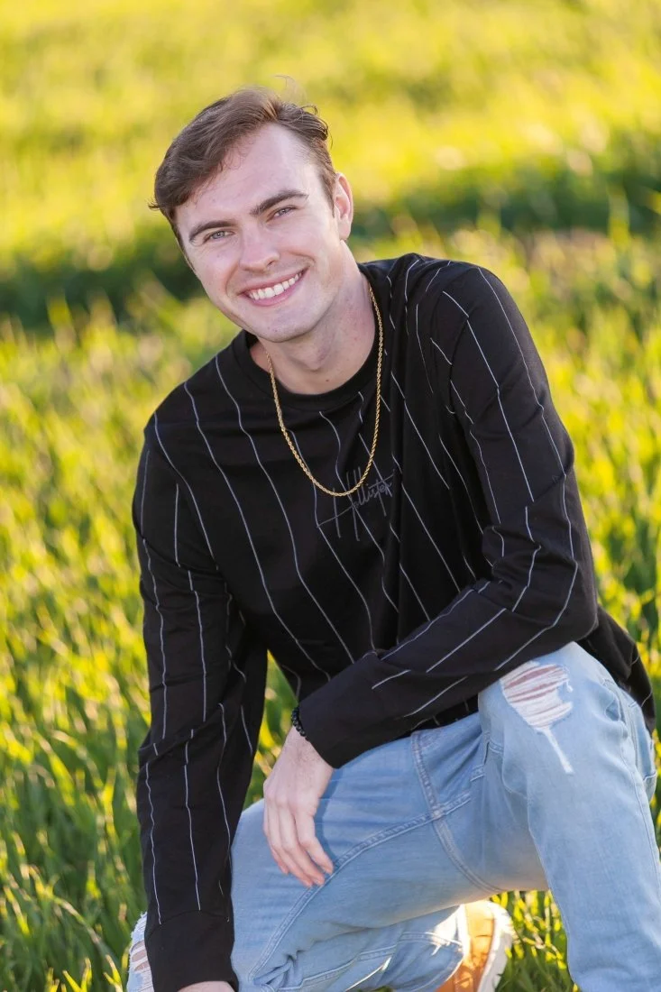 Young man smiling outdoors in a grassy field, wearing a black pinstripe shirt, ripped jeans, and a gold chain.