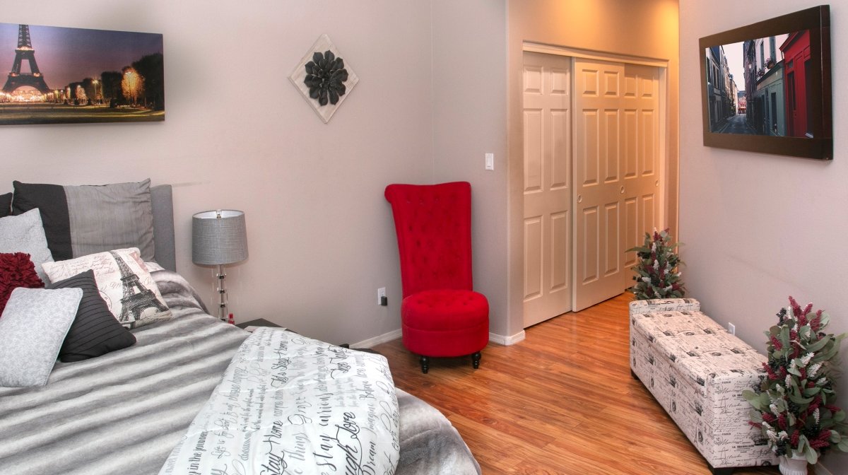 A bedroom with a bed, pillows, and a lamp on a nightstand. There is a red velvet chair, a closet with double doors, a framed city street photo on the wall, and small Christmas trees in the corner.