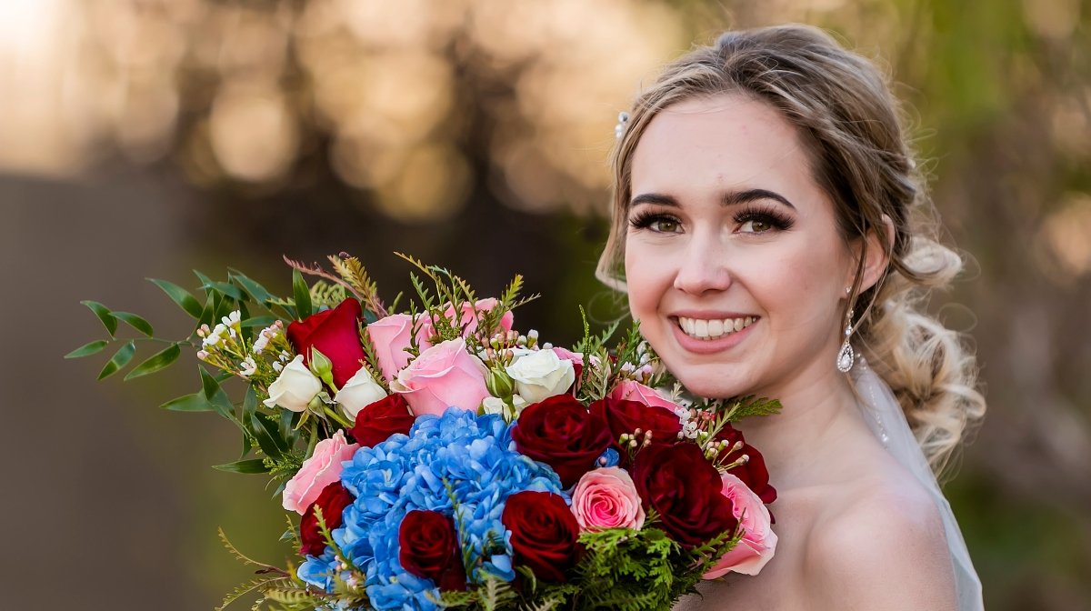 Brunette bride smiling and holding a colorful bouquet of roses and hydrangeas outdoors.