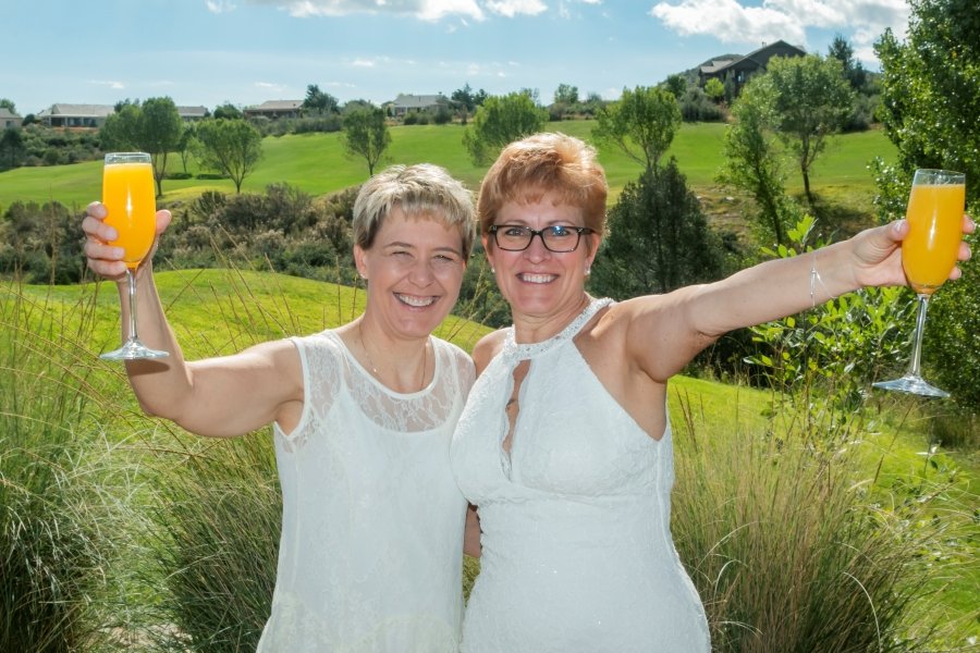 Two women in white dresses smiling, holding glasses of orange juice outdoors in a garden with green grass, trees, and houses in the background on a sunny day.