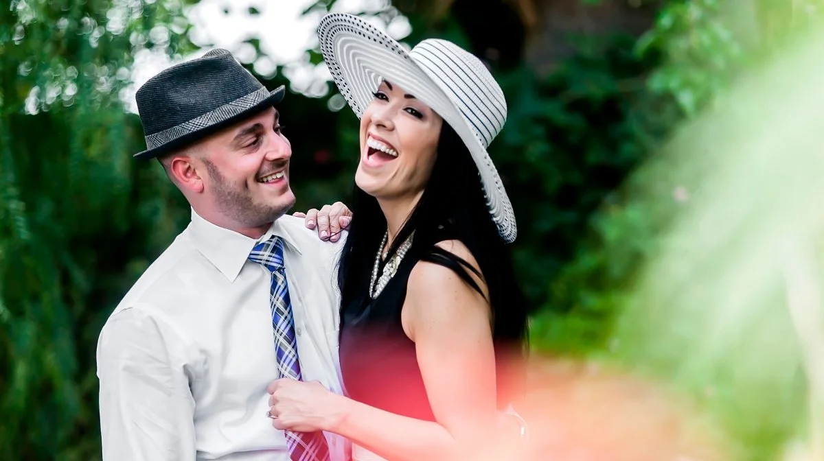 A smiling couple outdoors, the woman wearing a large striped sunhat and a black dress, and the man wearing a fedora hat and a tie.