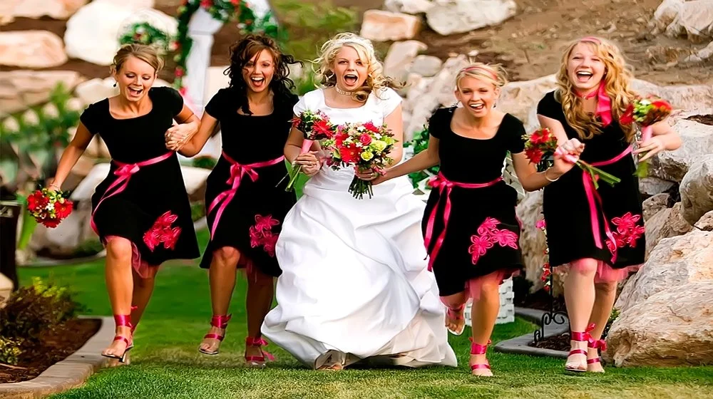 A bride in a white wedding dress holding a bouquet, walking with four bridesmaids in black dresses with pink accents, all smiling and holding smaller bouquets, on a grassy outdoor area with rocks and greenery.