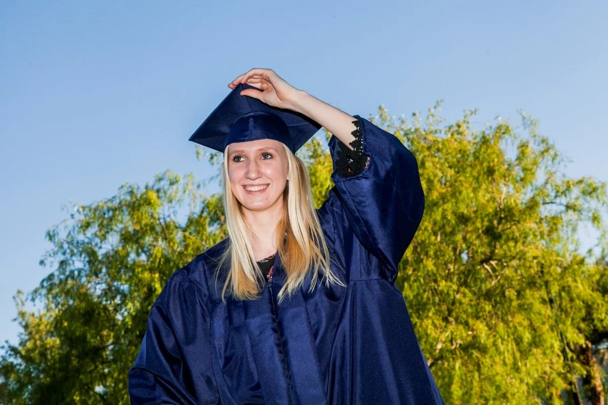 Young woman in a navy blue graduation gown and cap smiling outdoors, with green trees and a blue sky in the background.