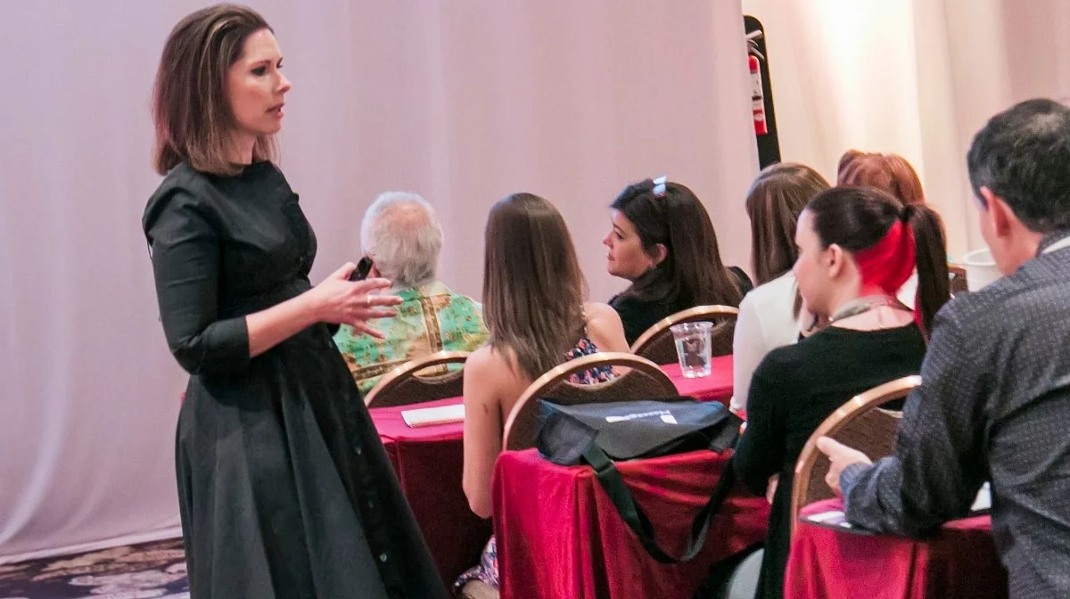 A woman standing and speaking to an audience of mostly women at a conference or seminar.
