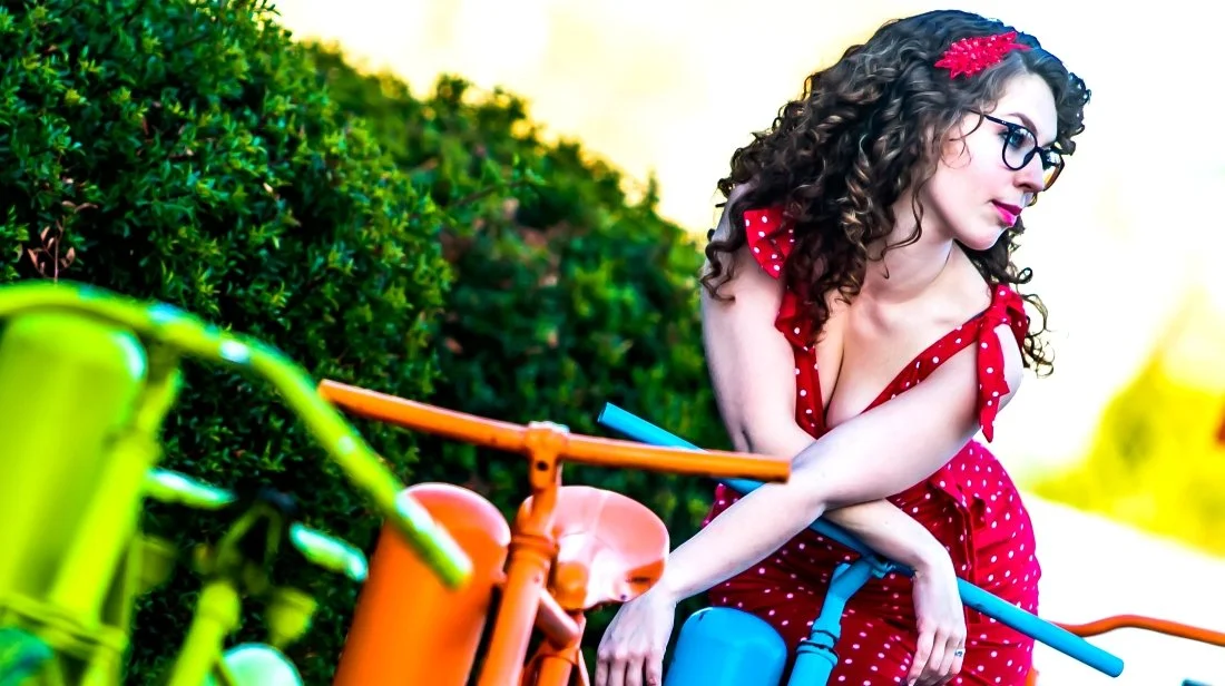 A woman with curly hair, glasses, and a red polka dot dress leaning on an orange bicycle, outdoors near green bushes.