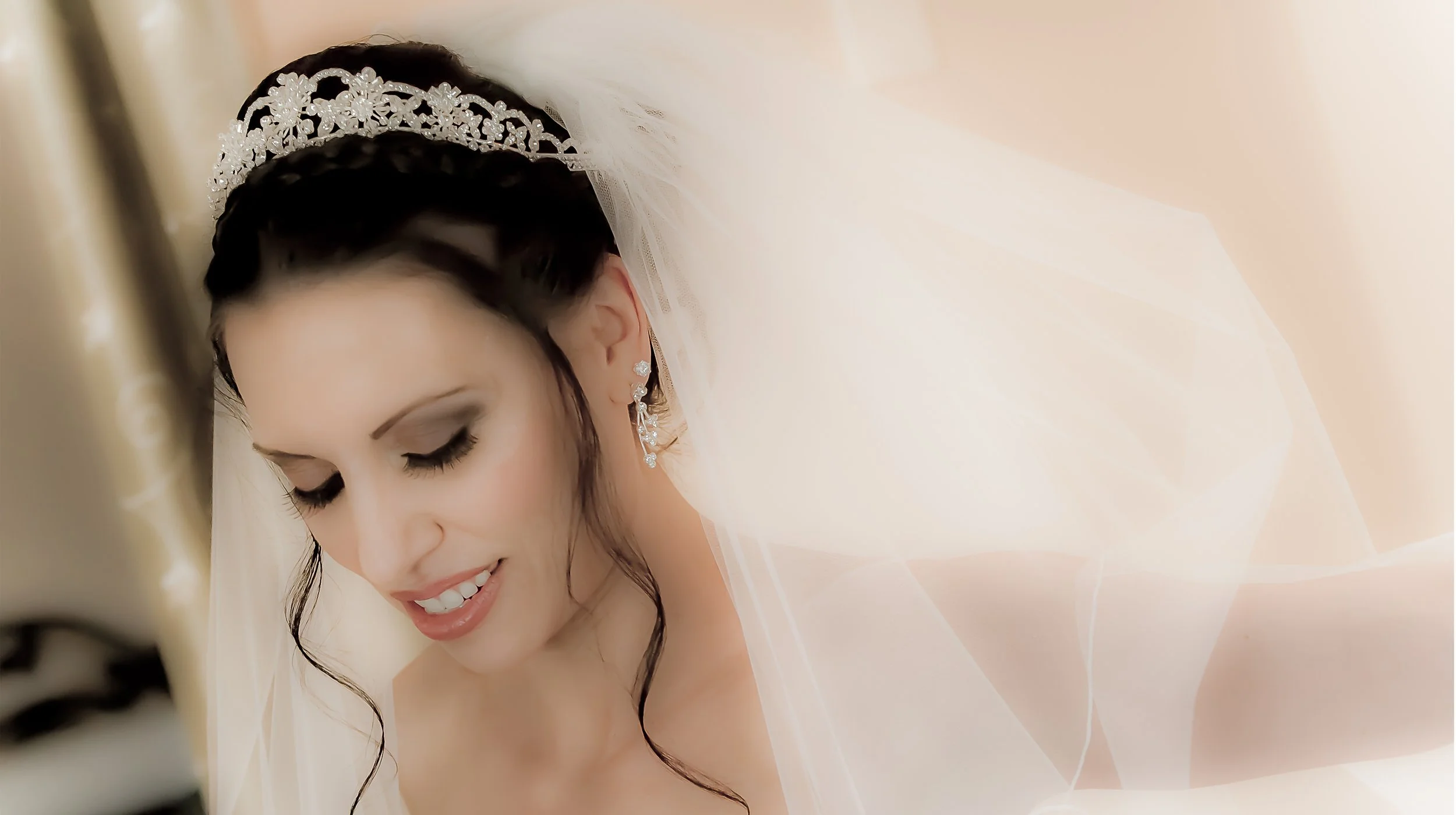 Close-up of a bride with dark hair wearing a tiara, earrings, and a veil, looking down with a gentle smile.