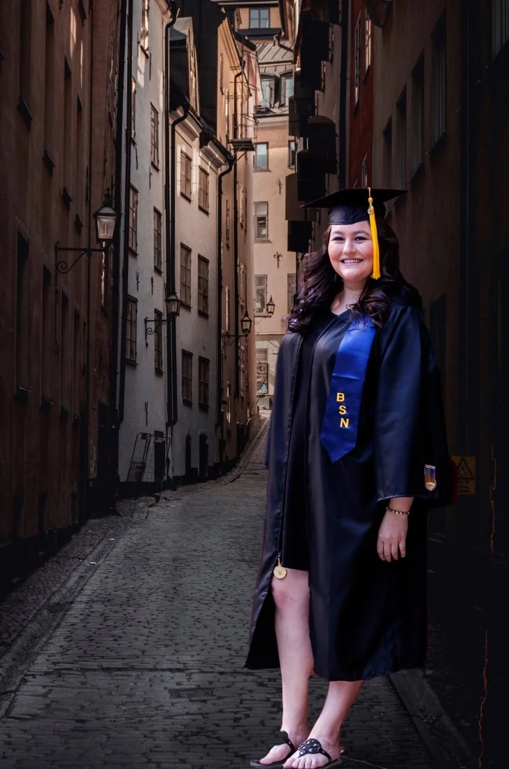 Young woman in graduation cap and gown standing on a cobblestone street lined with old buildings, smiling at the camera.