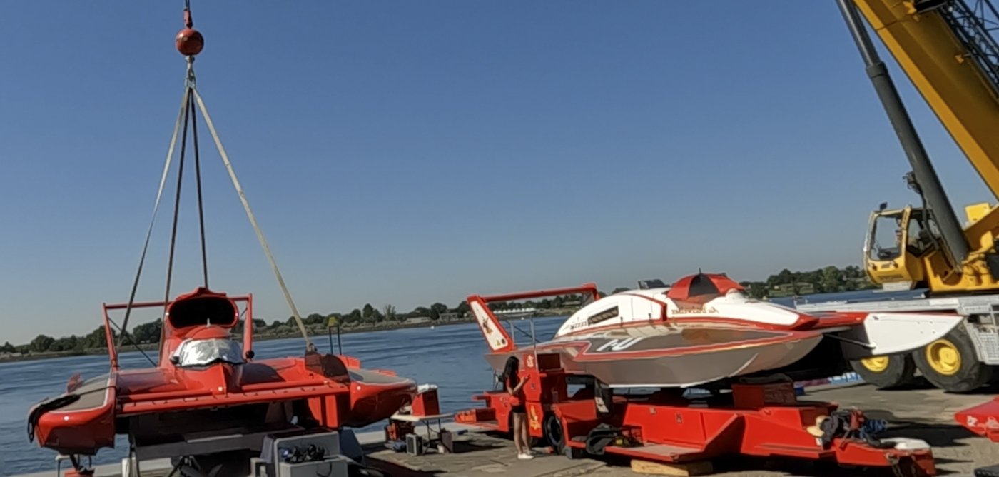 A racing boat being moved by a crane on a dock near the water, with another boat on a trailer nearby.