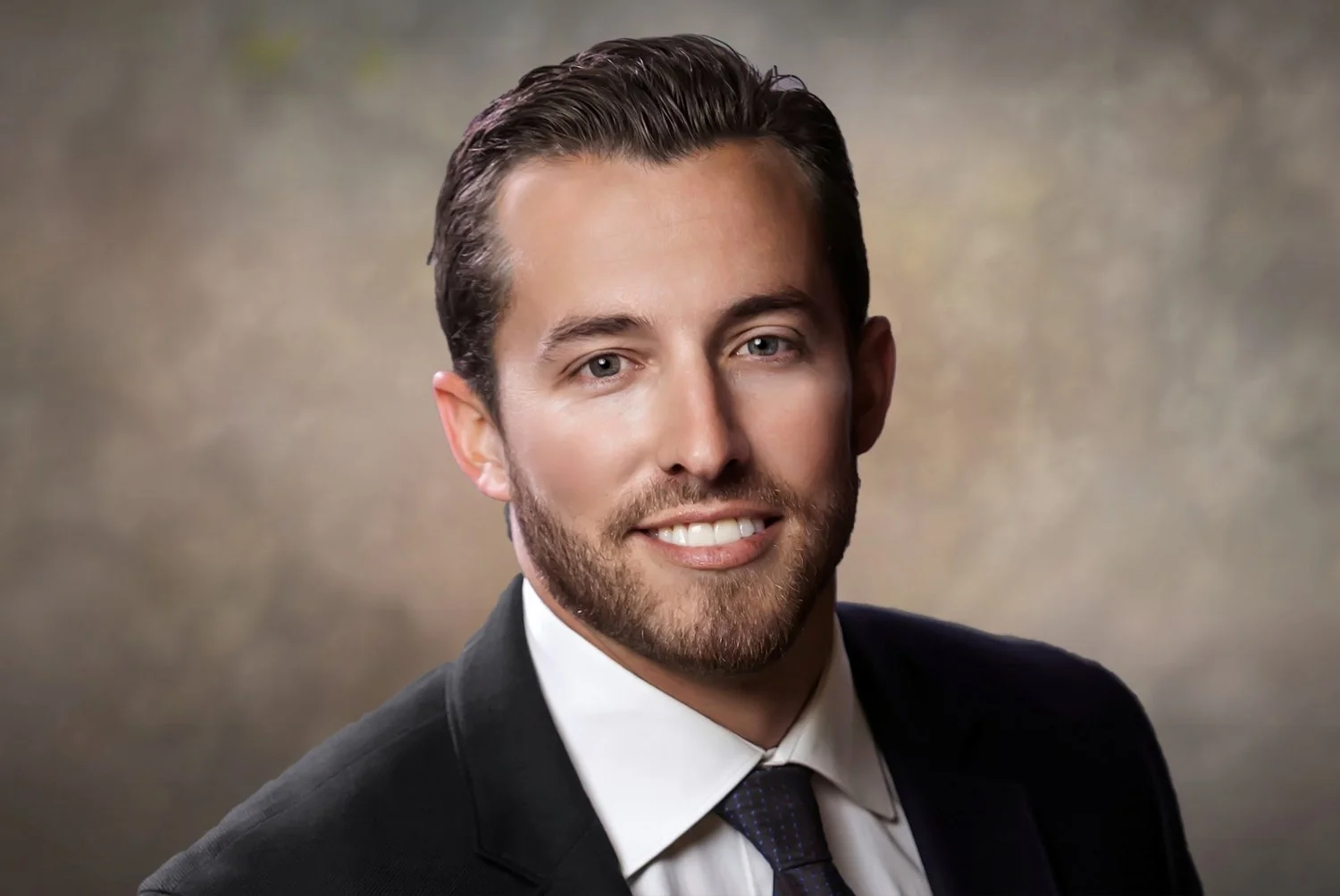 Headshot of a smiling man with dark hair, beard, wearing a dark suit, white shirt, and patterned tie, against a neutral background.
