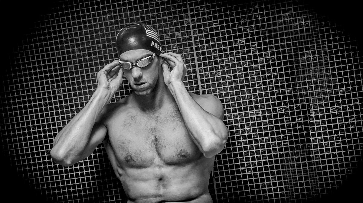 Male swimmer adjusting his goggles while standing shirtless in front of a tiled wall, wearing a swim cap and goggles.