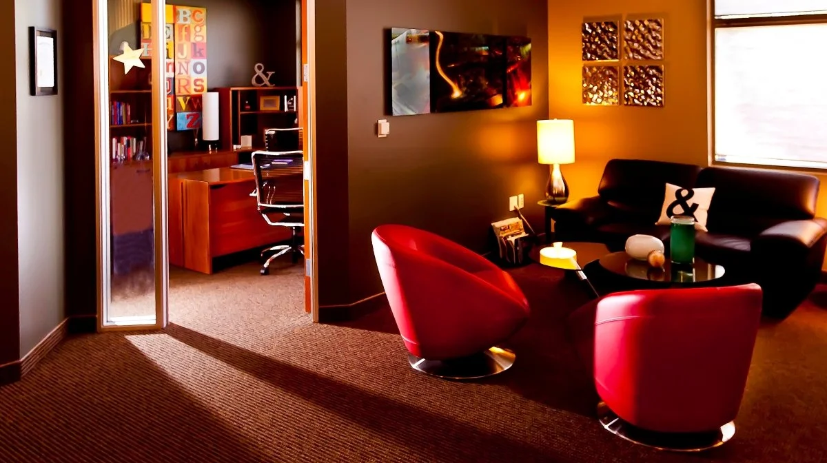 Living room with a black sofa, red chairs, and a side table with a lamp. A painting and decorative wall art are on the wall, and a window letting in natural light.