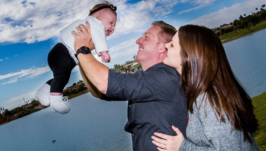 Family outdoors by a lake, father lifting a smiling baby girl, mother wrapping her arms around father, all enjoying a sunny day.
