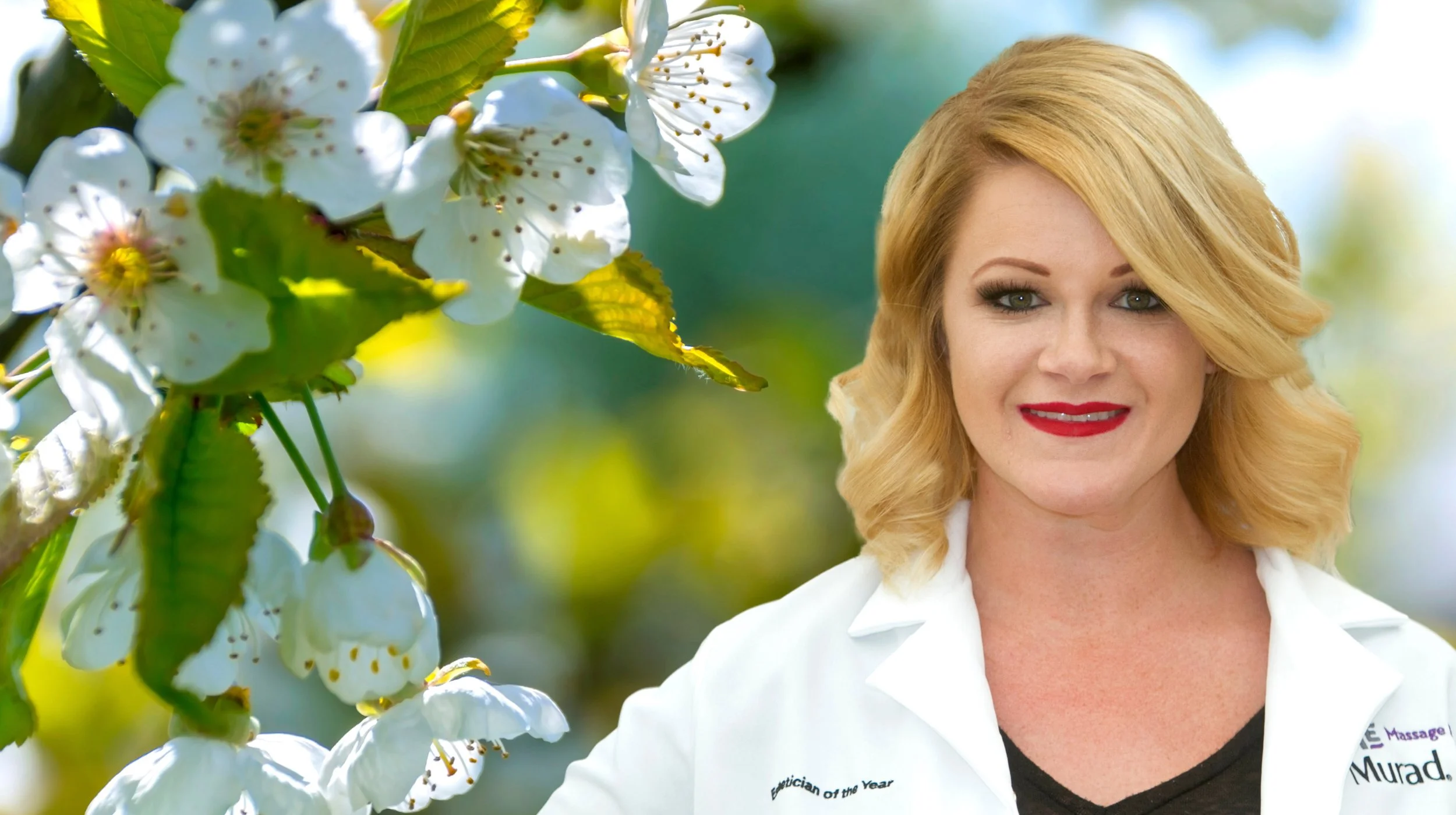 A woman with blonde hair and red lipstick smiling, positioned outdoors with blooming white flowers and green leaves in the background.