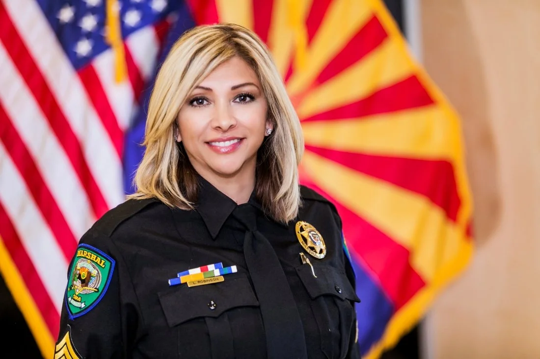 A smiling female police officer in uniform with American flags and a Enchantment, Arizona flag in the background.