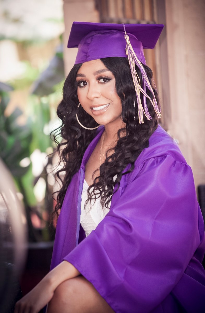 Young woman in a purple graduation cap and gown smiling, with long curly black hair and hoop earrings, sitting indoors with blurred plants in the background.