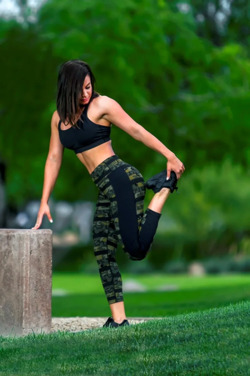 A woman in athletic wear stretching outdoors in a park with trees in the background.