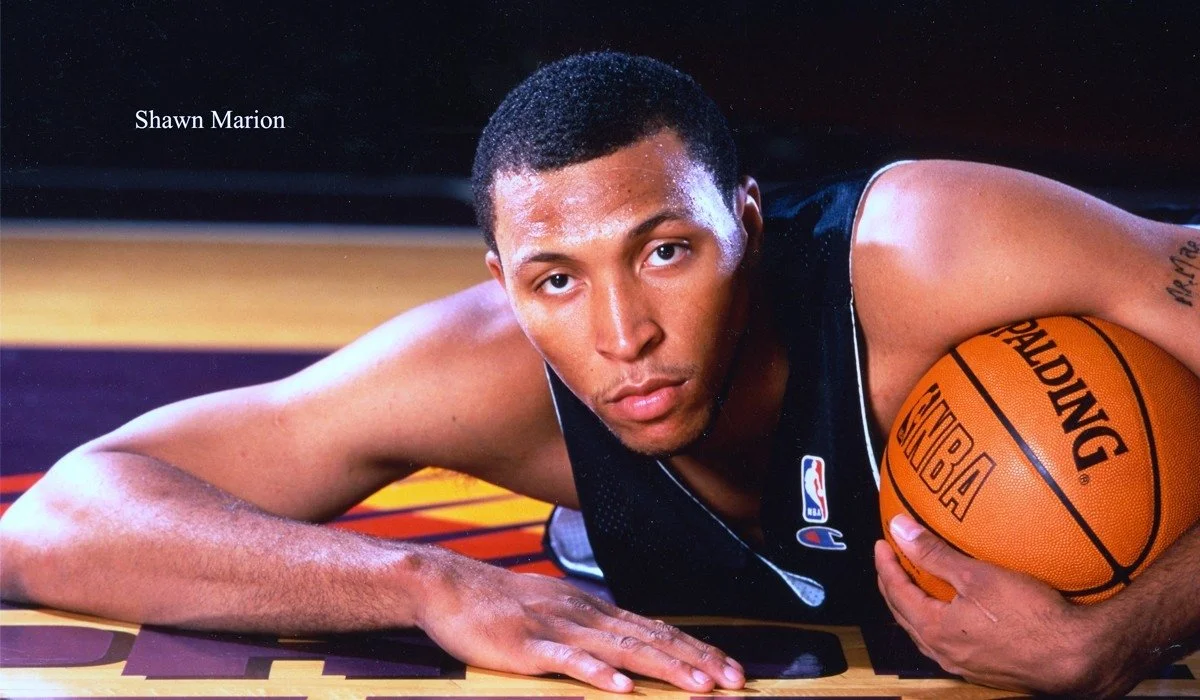 A young male basketball player lying on the court with a focused expression, holding a Spalding NBA basketball, wearing a black sports jersey with a small NBA logo.