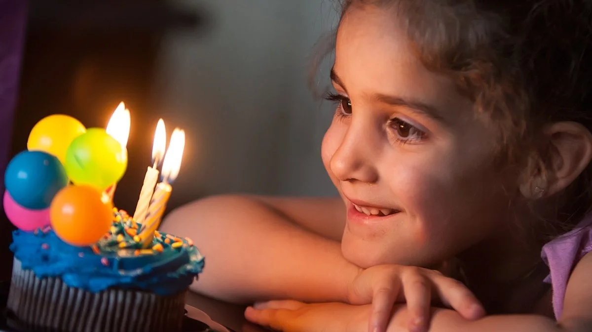 A young girl with curly hair looks at a birthday cake with lit candles and colorful balloons on top, smiling in anticipation.