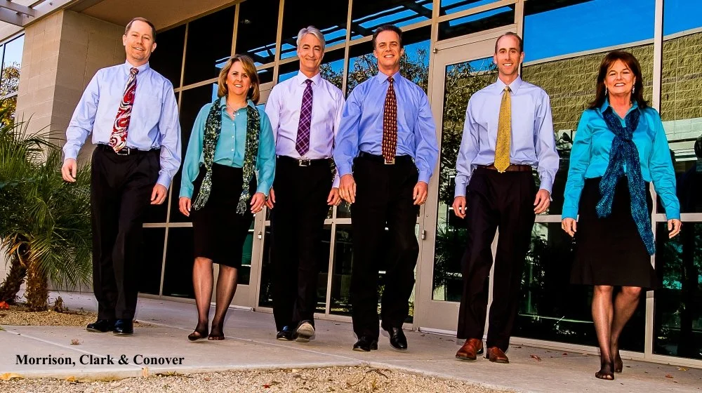 A group of six professionally dressed people standing outside a building with glass windows, smiling at the camera.