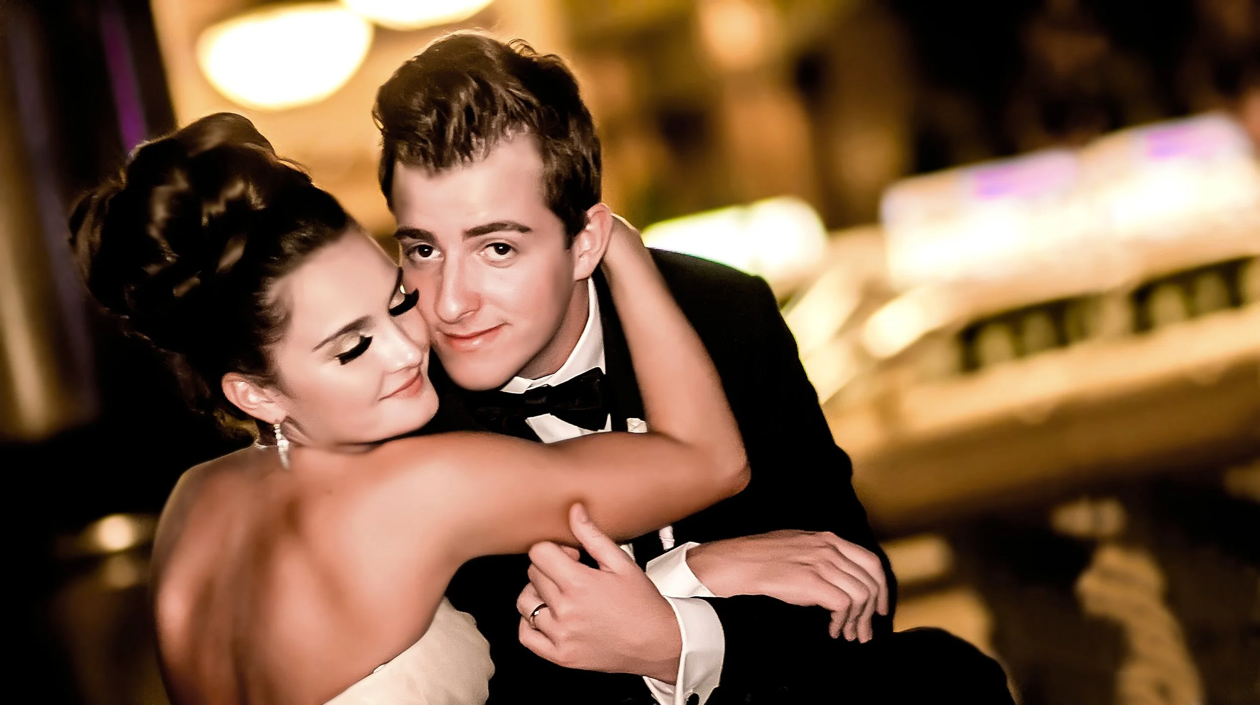 A young couple in formal attire embraces each other at a glamorous event, with warm lighting and blurred background.