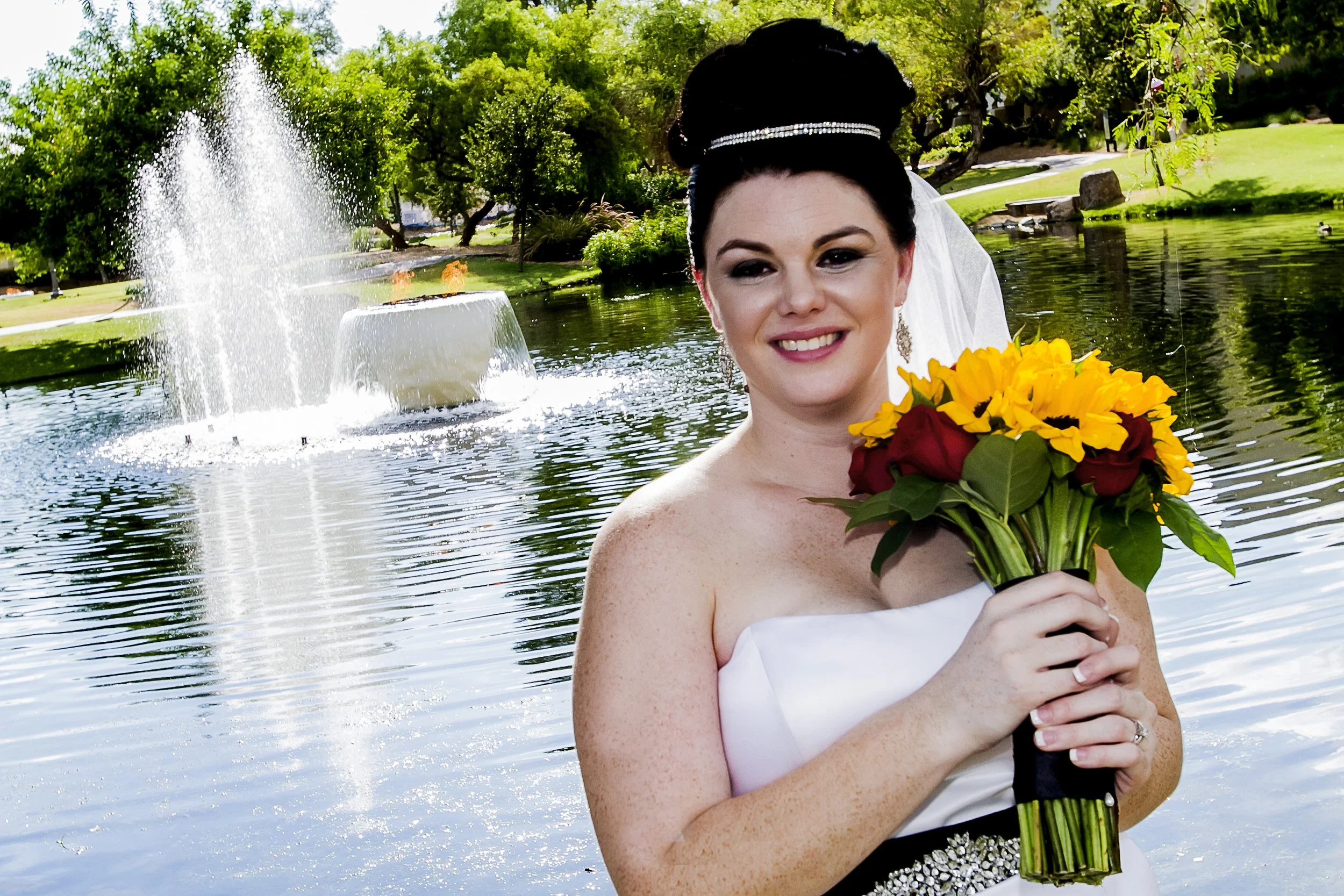 A bride with dark hair styled up, wearing a white strapless wedding dress with a jeweled waistband, holding a bouquet of yellow sunflowers and red roses, standing by a pond with a fountain and surrounded by green trees and grass.