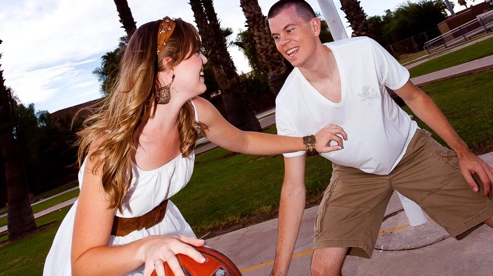 A woman and a man playing basketball outdoors on a sunny day, surrounded by palm trees.