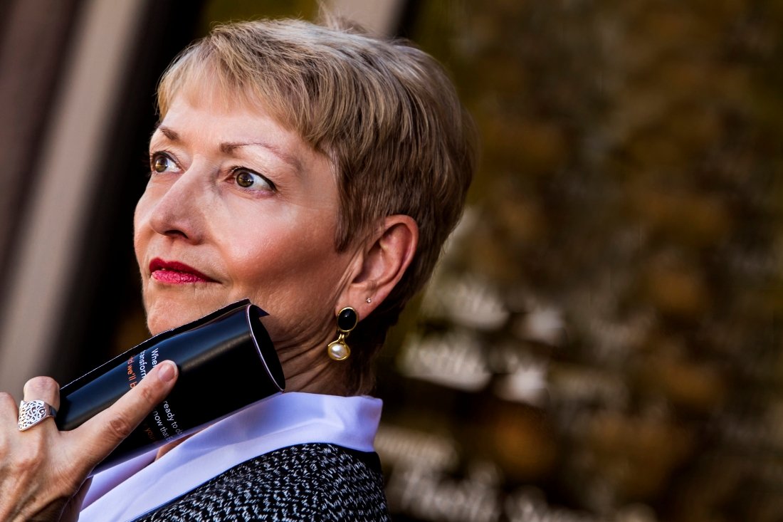 A woman with short blonde hair and pearl earrings is holding a black cylindrical object near her face, with a blurred outdoor background.