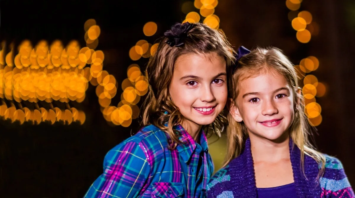 Two young girls with curly hair smiling outdoors at night, with blurred string lights in the background.