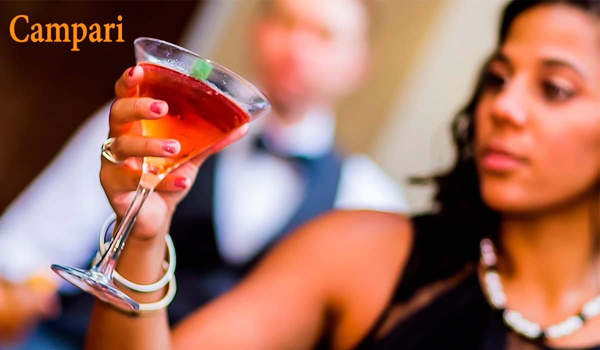 A woman holding a glass of red cocktail with a lime wedge, with a blurred woman and man in the background.
