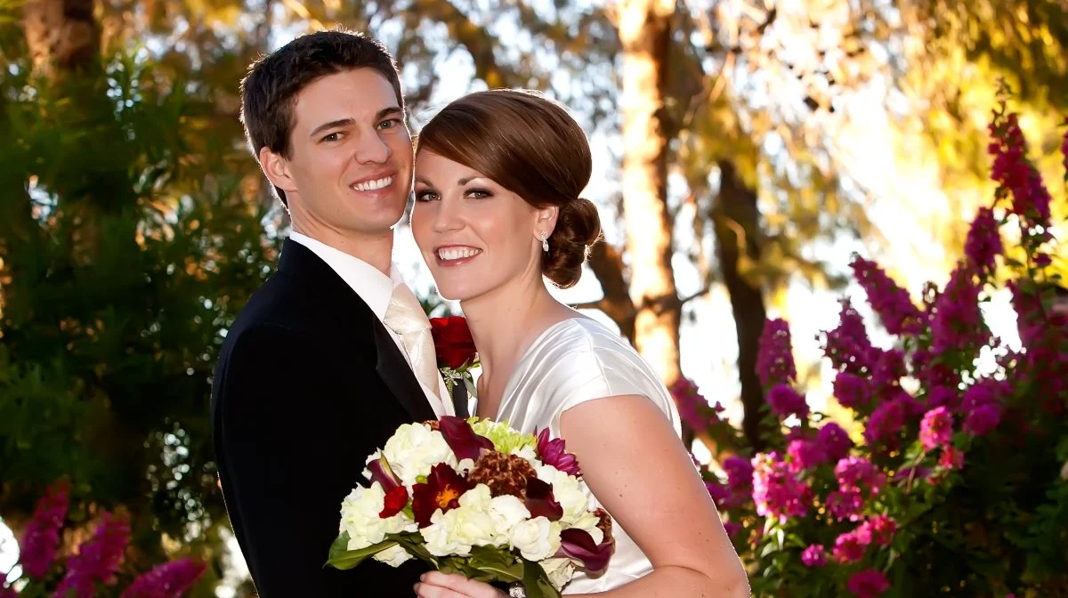 A smiling young couple at their wedding, standing outdoors with trees and pink flowers in the background. The bride is holding a bouquet of white, red, and purple flowers.