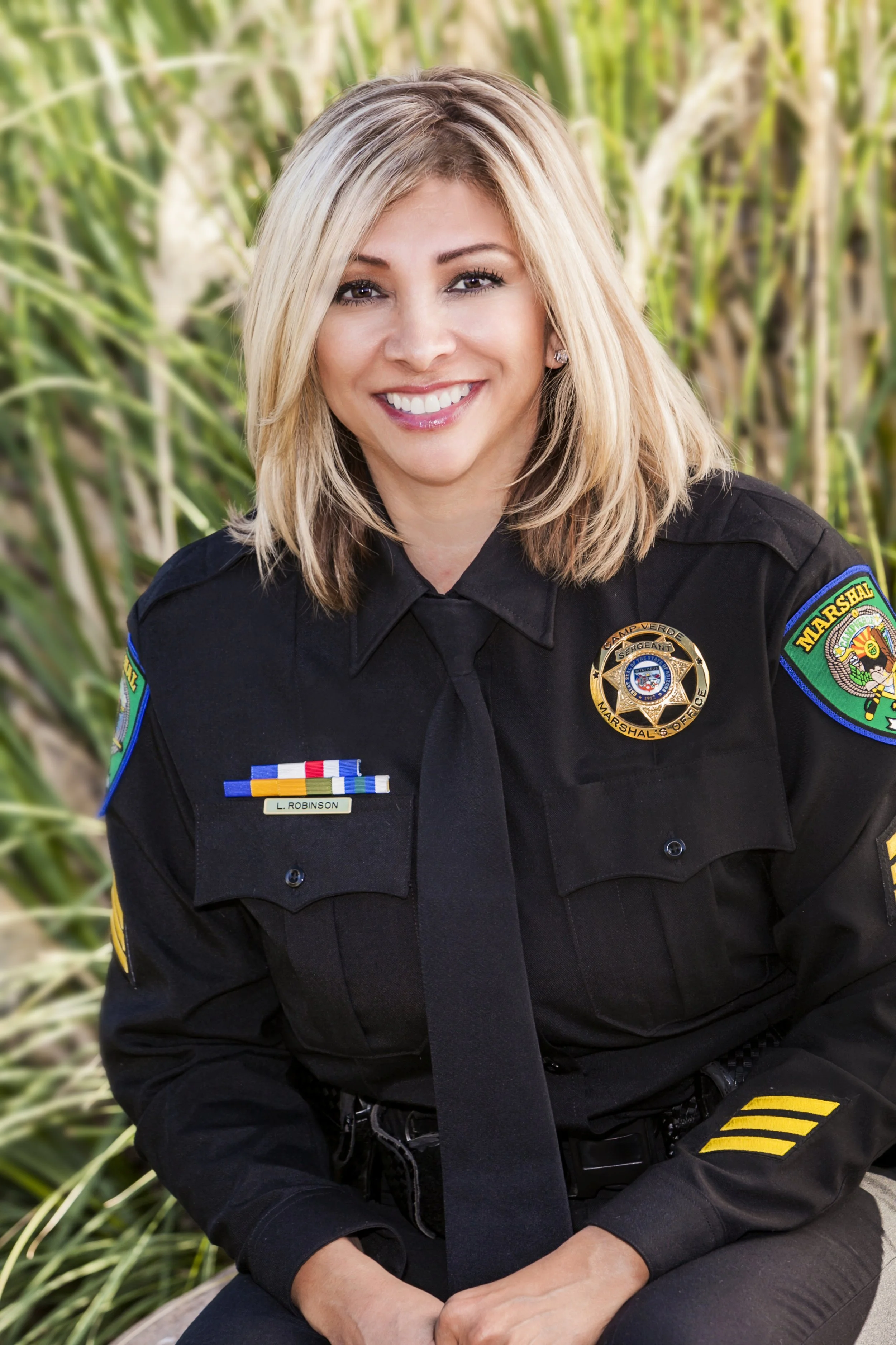 A smiling female police officer in uniform sitting outdoors with greenery in the background.