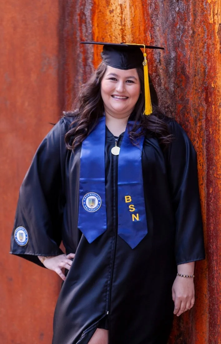 A young woman in a graduation cap and gown, standing against a rust-colored wall, smiling with her hand on her hip.