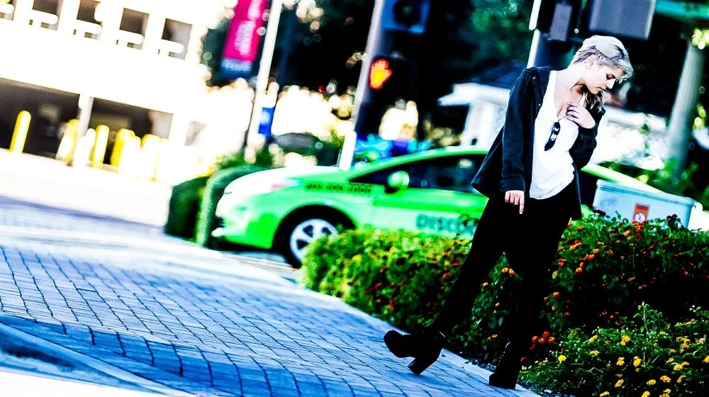 A woman with short blonde hair, dressed in black and white, walking on a city sidewalk next to a flowerbed, with a green taxi in the background.